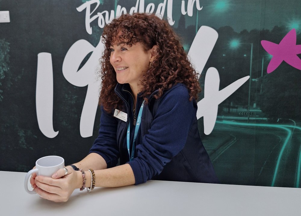Person seated at a table holding a cup with both hands, wearing a dark long-sleeve top and a lanyard. The background features a dark wall with large white text that reads ‘Founded in 19’ and a pink star graphic partially visible.