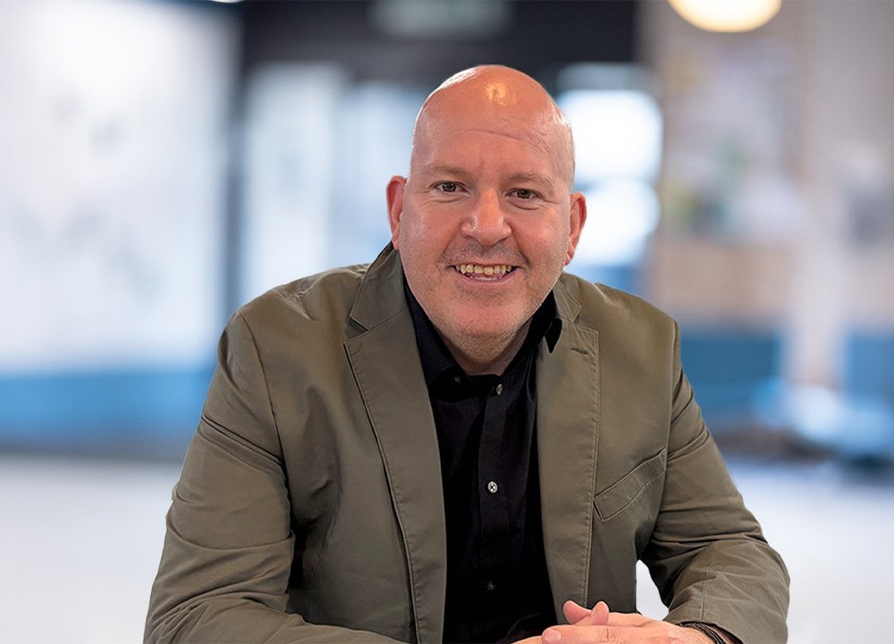 Professional headshot of a person in a blazer seated indoors with a blurred office background.