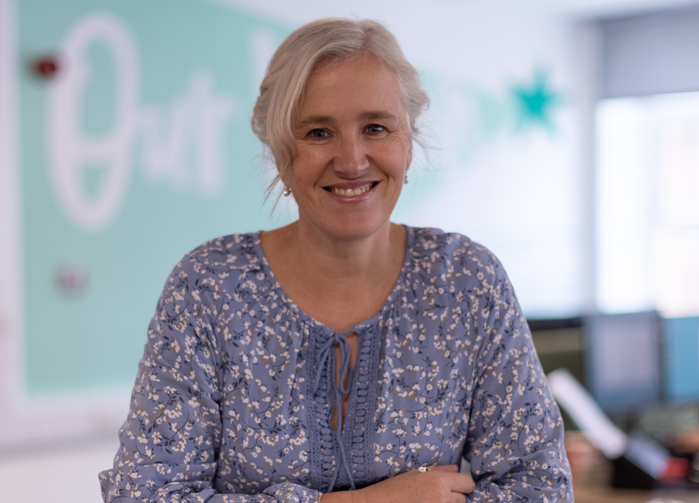 Woman with light hair in a blue floral blouse smiling in an office setting.