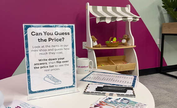 A tabletop display sign reads “Can You Guess the Price?” beside a mug, pens, and clipboards. Behind it stands a small wooden toy stall with a striped canopy and miniature items like a teddy bear and fruit basket. 