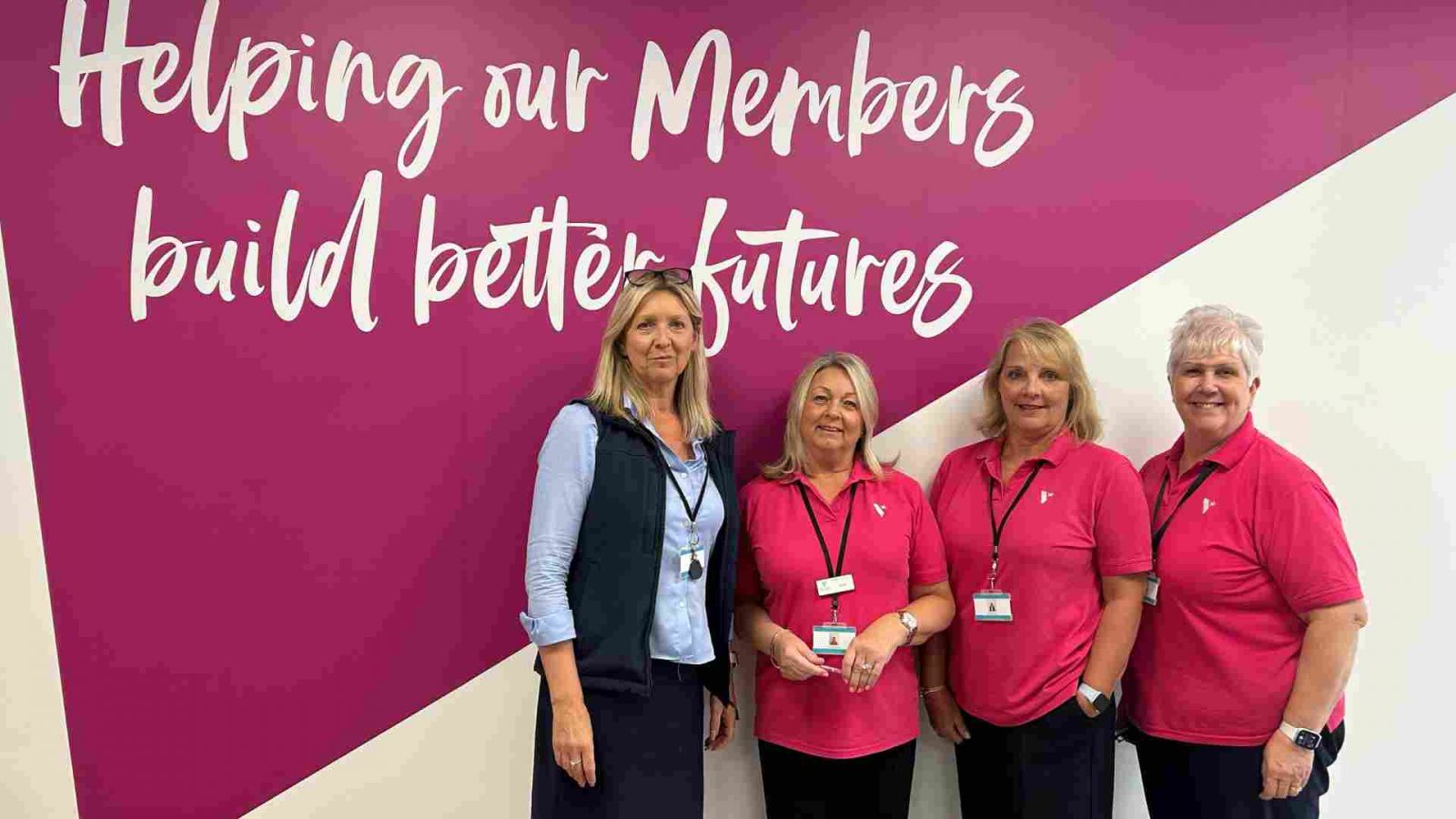 Four staff members standing in front of a pink wall with white text that reads ‘Helping our Members build better futures.