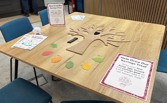 A wooden table displays a savings‑themed activity called “Help Grow Our Savings Tree.” A large wooden cutout of a tree lies flat on the table, surrounded by colourful paper leaves and marker pens. 