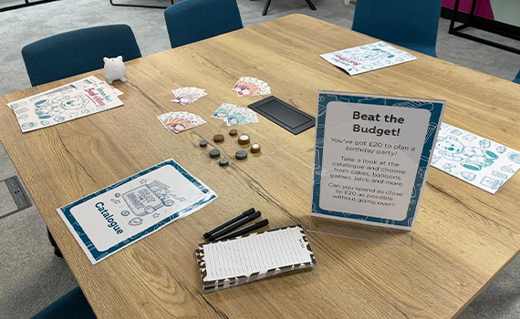 A wooden table is set up for a fun budgeting activity called “Beat the Budget.” A sign explains the challenge: plan a birthday party with £20 using a catalogue of items. 