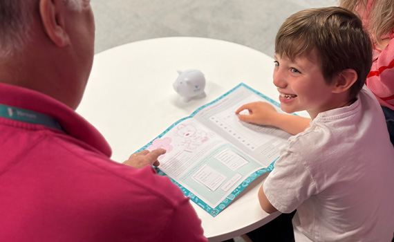 A smiling young boy sits at a round white table with two adults, looking at a colourful savings activity sheet.